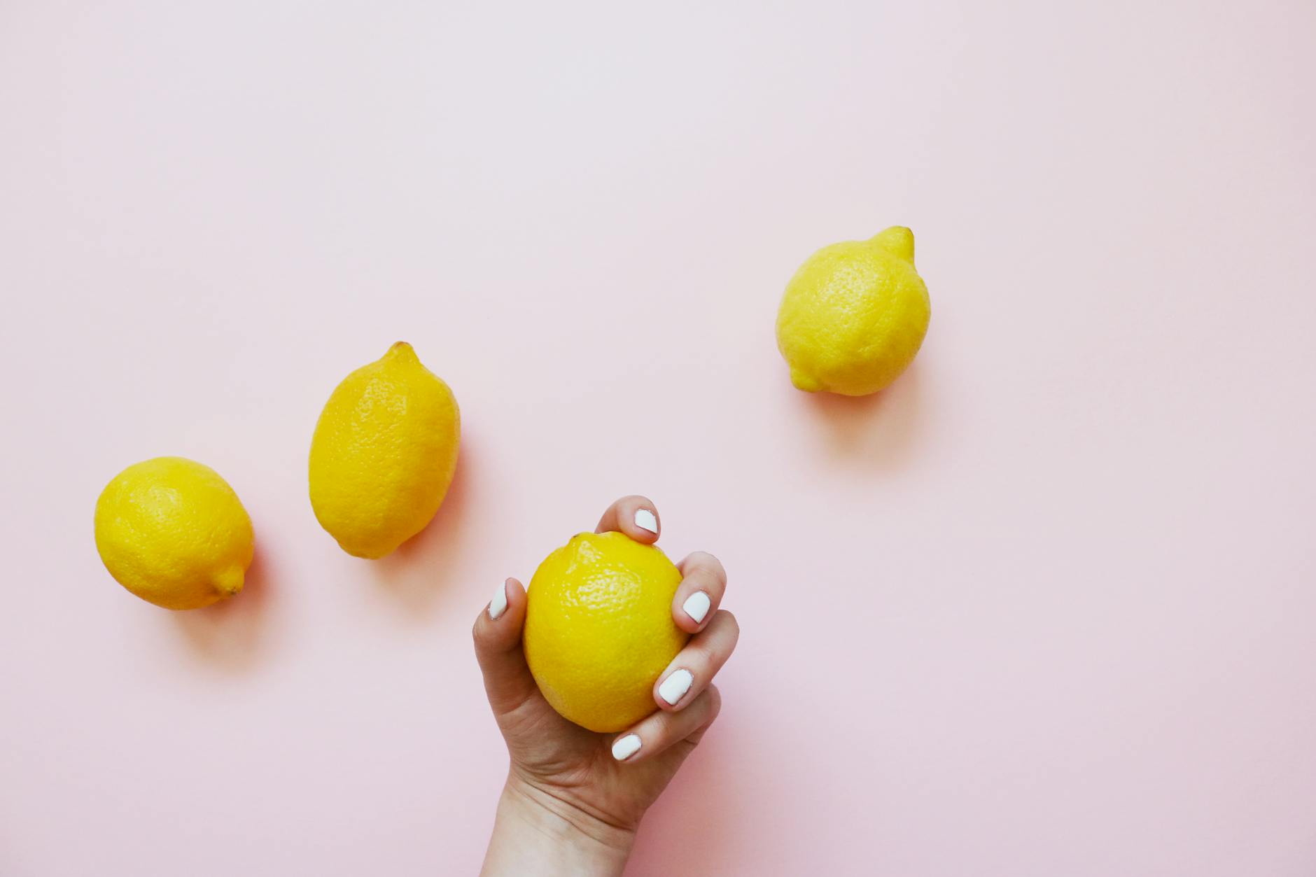 A hand holding a lemon on a soft pink background with additional lemons nearby