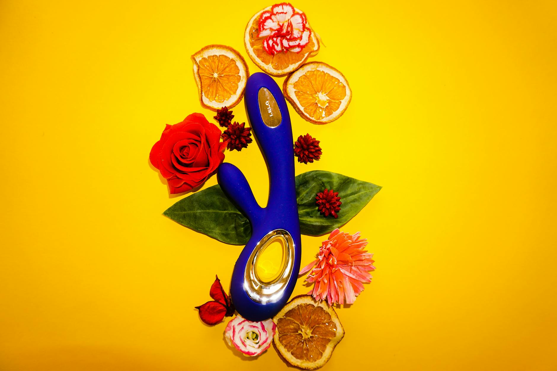 Vibrador colorido rodeado de flores y rodajas de naranja sobre fondo amarillo